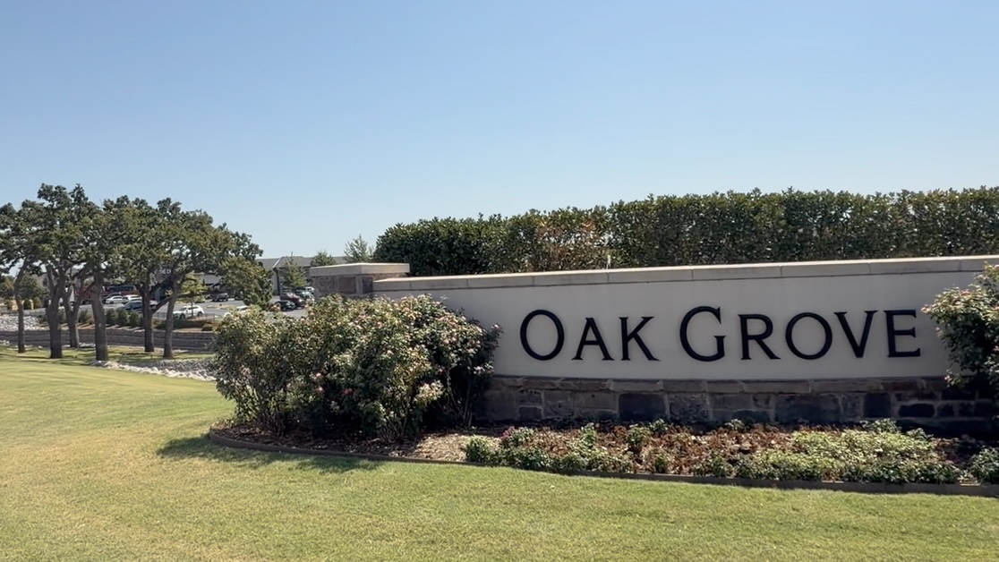 Establishing photograph of the OAK GROVE entrance sign with the mature oak row visible beyond or beside it; frame the lettering clearly while keeping enough of the trees in the composition to create the visual irony between the place name and the grove's altered crowns.
